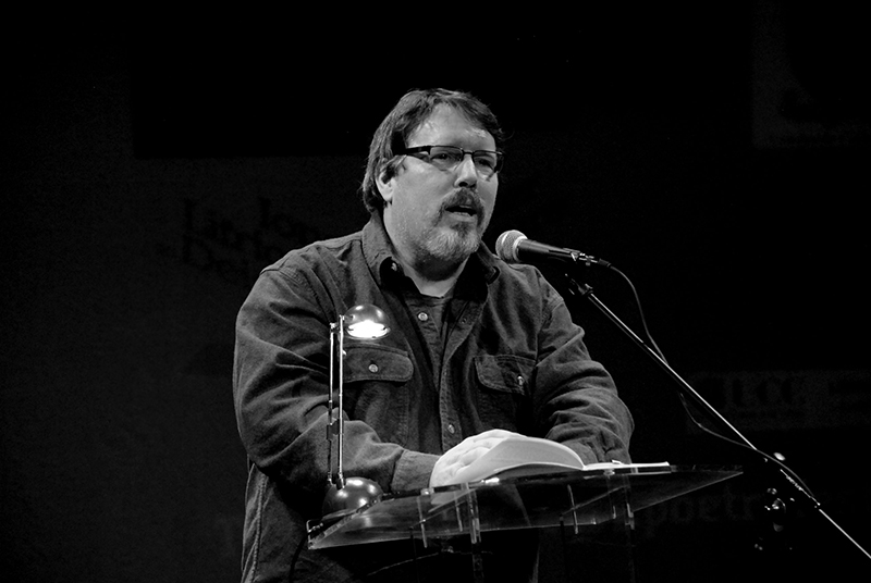 Black and white photo of Brian Turner reading from a book on a lectern. He is speaking into a microphone.