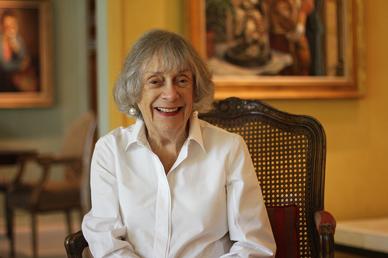 Mary Hyman seated in a chair in her home, smiling, with artwork hanging in the background.