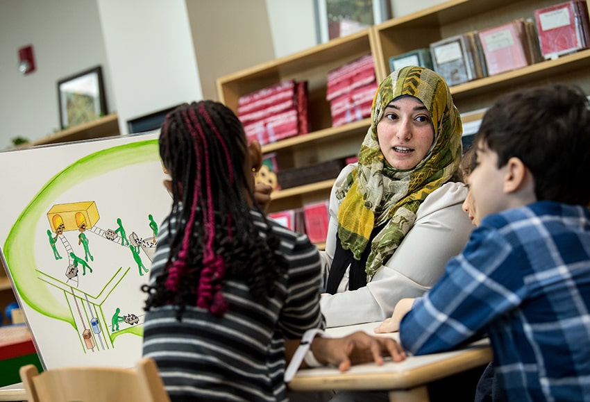Female teacher wearing hijab sits with two students explaining a poster diagram