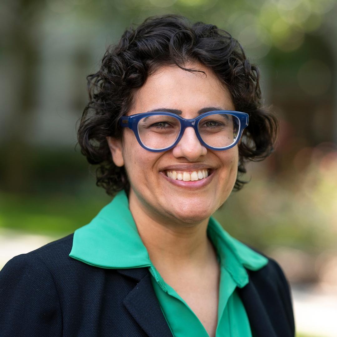 Marina smiles in a green shirt and black blazer, wearing blue framed eyeglasses with Loyola