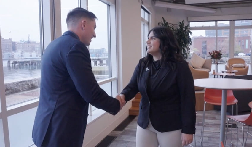 A female Emerging Leaders MBA student with a dark blazer with a Loyola “L” pin on her lapel shakes hands with an employee at Brown Advisory