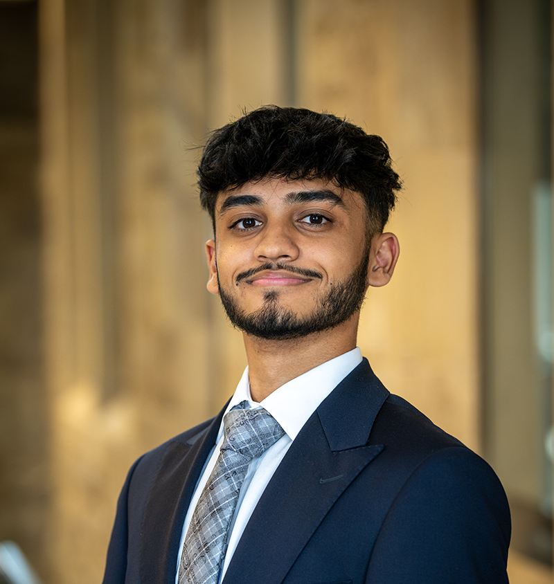 A young man with short, dark hair and a neatly trimmed beard wears a navy suit, light blue shirt, and patterned gray tie, posing confidently with a slight smile against a softly lit indoor background.