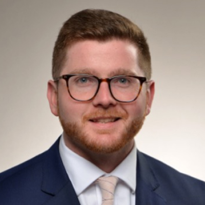 Professional headshot of a Devon De Marco depicting a young man with glasses and a neatly trimmed beard, wearing a navy suit and light tie against a soft neutral background.