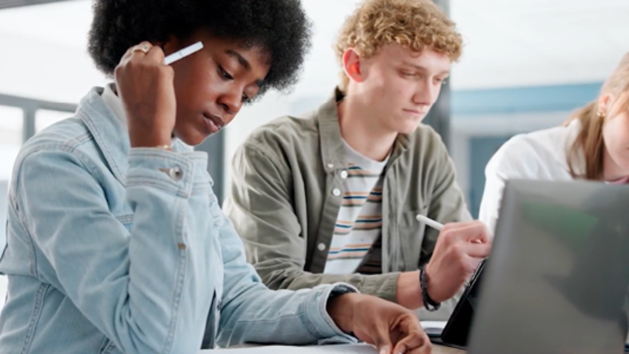 A focused woman in a denim jacket holds a pen, studying alongside a man in a striped shirt at a desk. - Press enter to play