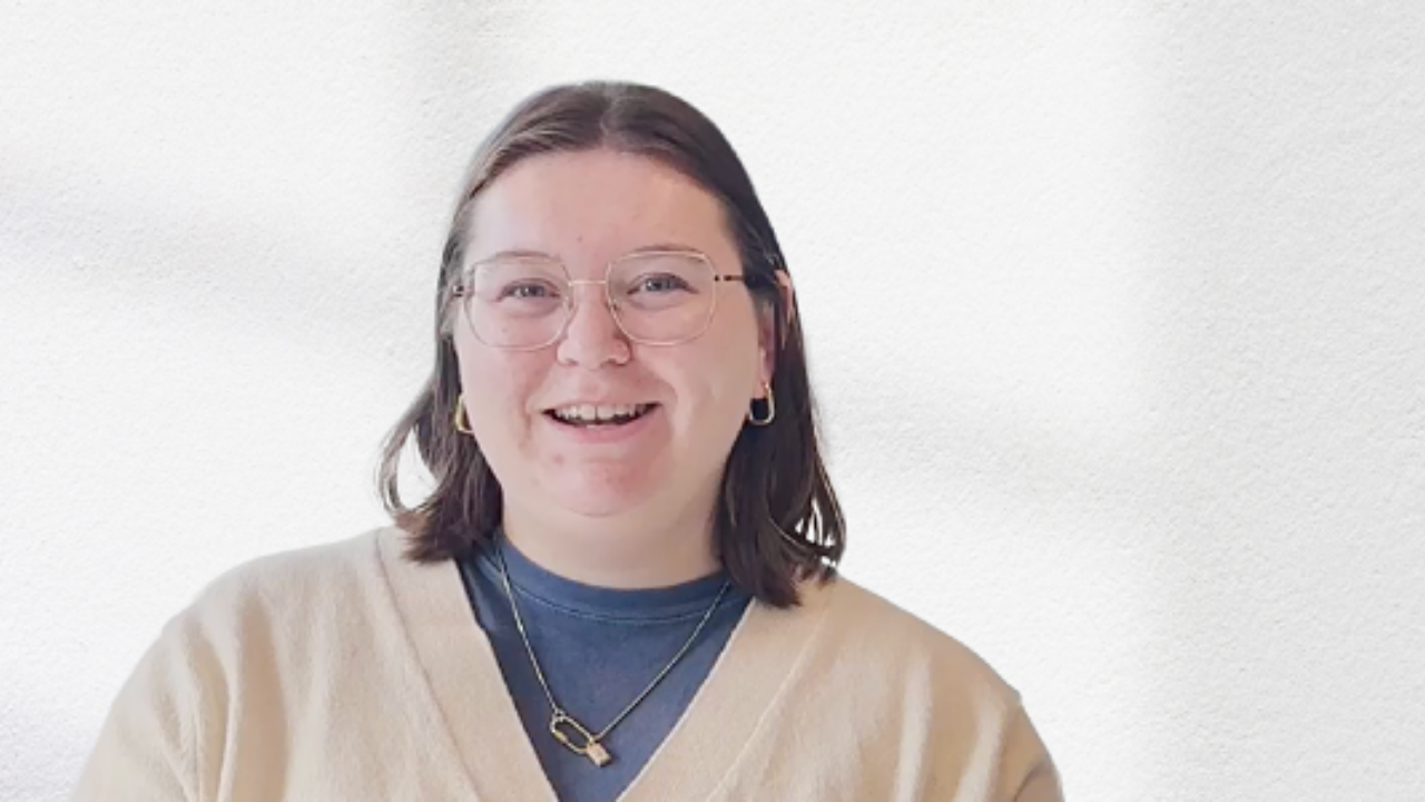 Smiling person with glasses and dark hair, wearing a beige sweater and blue shirt, stands indoors. - Press enter to play
