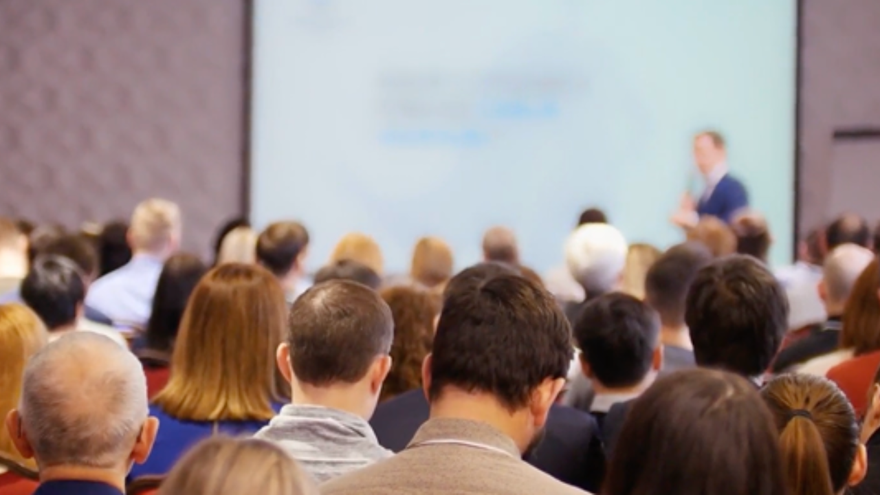 Audience attending a conference, viewing a speaker at a podium. The crowd is focused, seated in rows, highlighting an educational setting. - Press enter to play