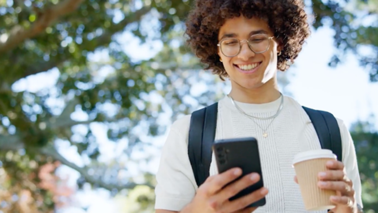 Smiling young man with curly hair and glasses, wearing a white shirt and backpack, holds a smartphone and coffee cup outdoors under sunny trees. - Press enter to play