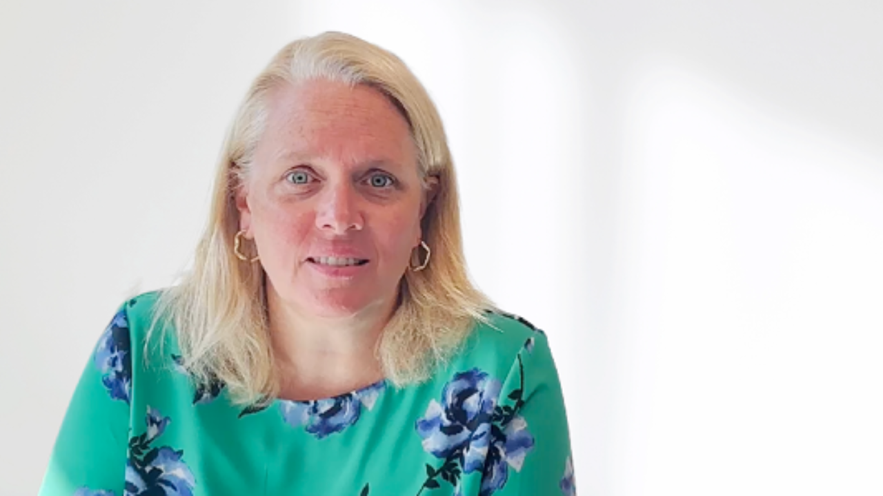 Woman with blonde hair wearing a floral blouse sits in an office. She has a friendly, professional expression. - Press enter to play