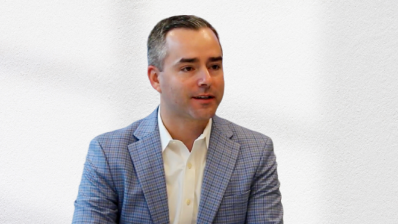 A man in a suit sits in a meeting room, speaking. - Press enter to play