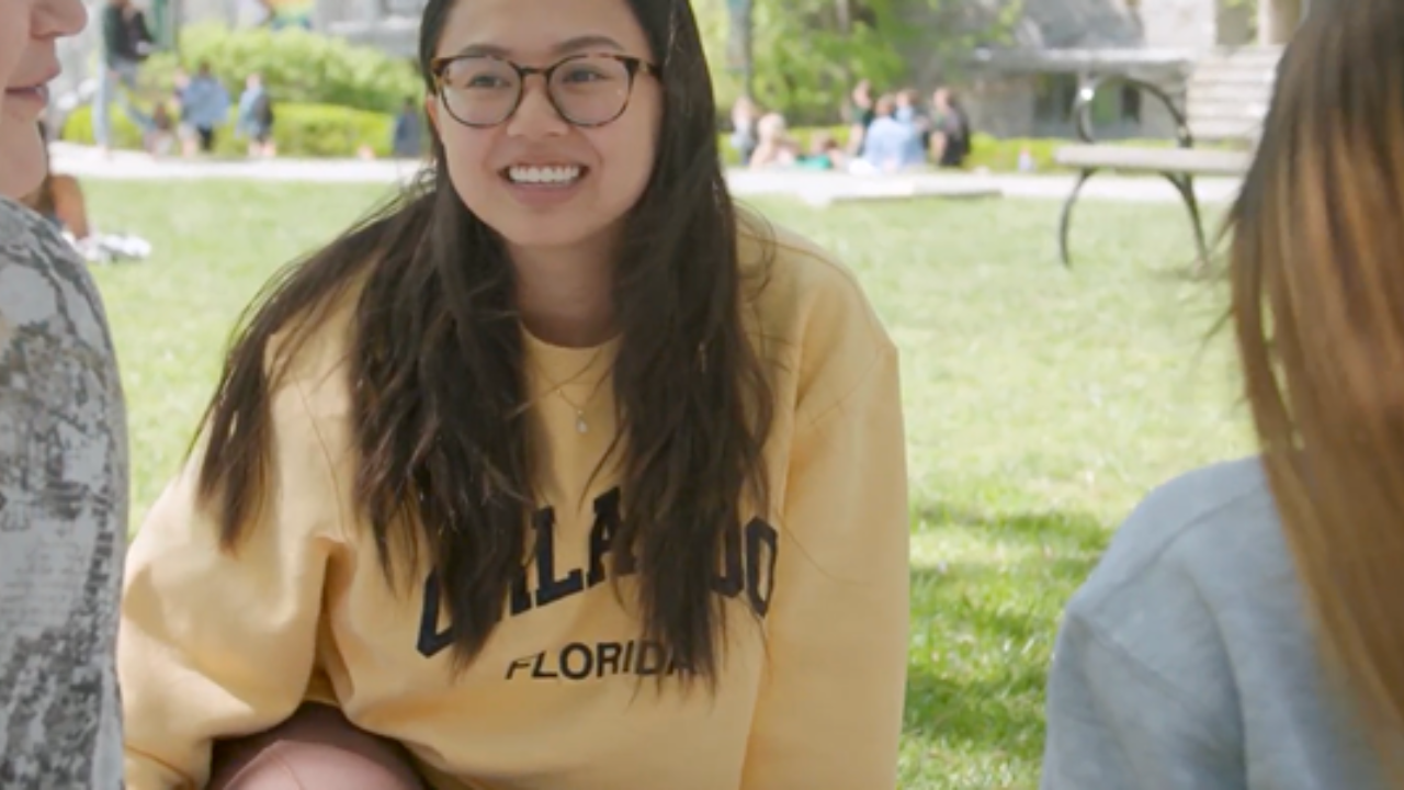 A young woman with glasses and a yellow sweatshirt smiles while sitting on grass. - Press enter to play