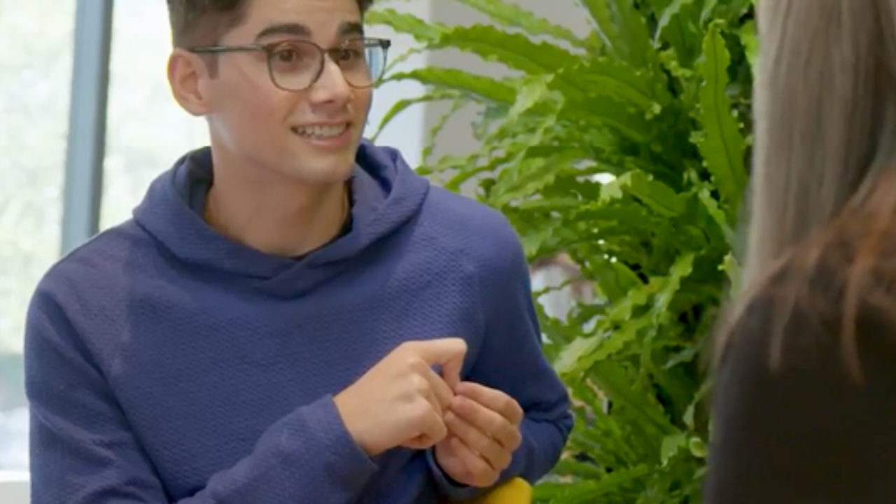 Young man in glasses and blue sweater smiles while speaking to a professor. - Press enter to play